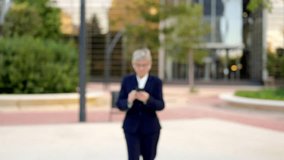 Horizontal video of a distracted mature businesswoman using phone approaching to the camera and smiling outdoors