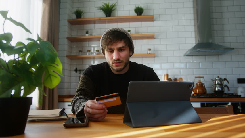 Young man with beanie sitting in kitchen at home entering credit card number on tablet device for makes distant goods purchase. Credit card is declined or out of limit, feeling sad and unhappy Royalty-Free Stock Footage #3411105137