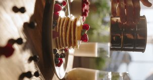 Unrecognizable woman sprinkles powdered sugar on perfect stack of pancakes, aesthetically decorated with berries, in slow motion, in kitchen scene. Cinematic vertical video. Beauty of tasty treats. 