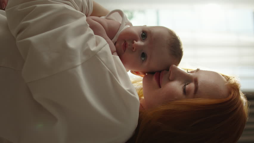 Close-up of cute baby holding onto his mom's shoulder radiates peace and serenity of motherhood, beautiful concept of parenthood. Cinematic advertisement, warmth and love childhood, vertical video