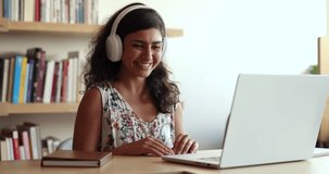 Laughing young 30s Indian woman in headphones make videocall, greeting colleague or mate start virtual meeting, educational webinar event sit at desk use laptop. Professional counseling, communication