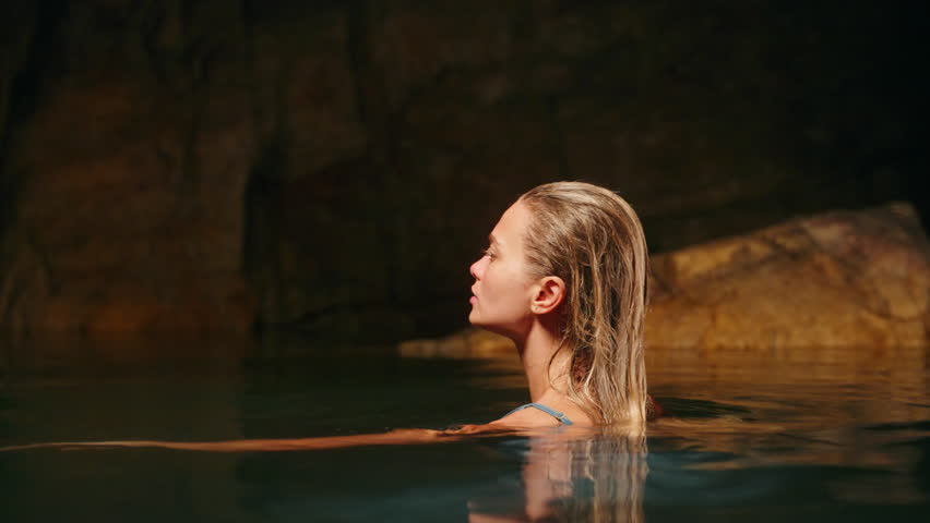 Ethereal woman in blue gown bathes at sea cove under moonlight. Mystic, serene female enjoys night swim, solitude. Fashion, skincare in nature scene. Rocks, water shimmer, cinematic beauty shot.