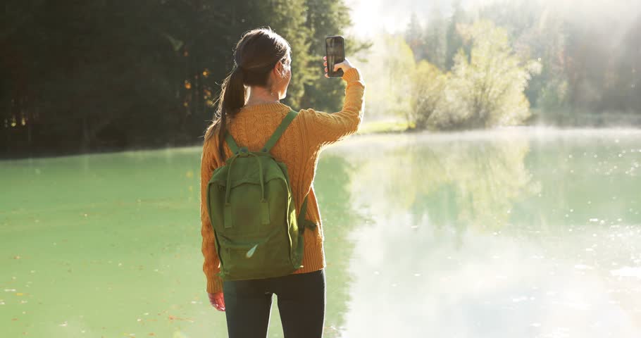 Young woman with a backpack shooting video with smartphone in the Alps. Back of young woman at an alpine lake. Sunny autumn day in the Alps. Social media creation concept.