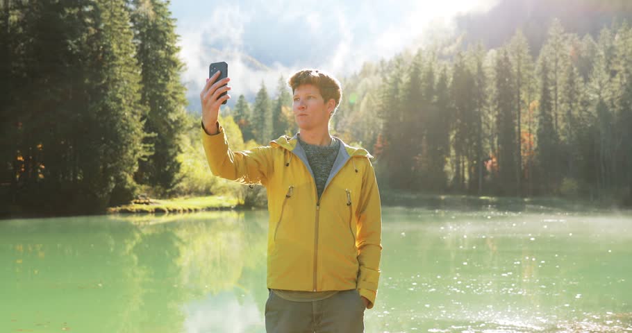 Young man at a scenic alpine lake in Jezersko, Slovenia. Male influencer talking to camera on smarphone. Young traveller capturing video footage on a trip.