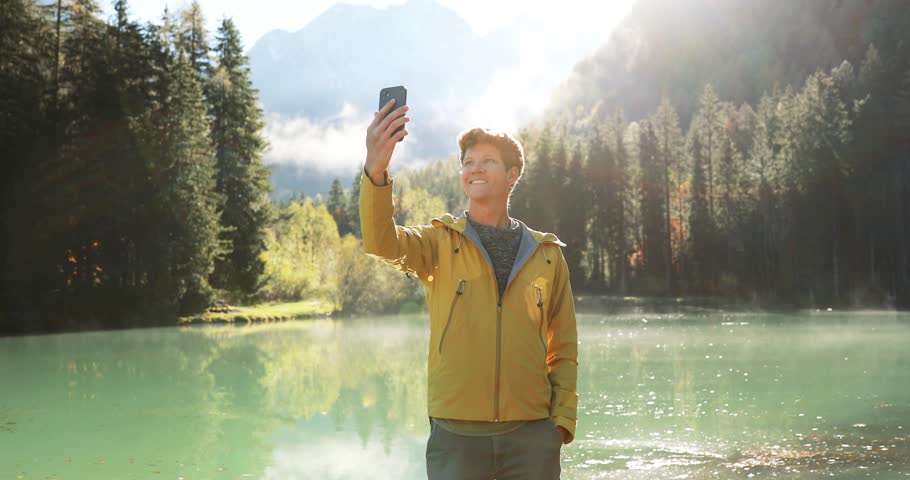 Young hiker in vibrant yellow jacket shooting video with smartphone. Smiling athletic young man capturing beautiful scenery at an alpine lake. Sunny autumn day in the Alps.