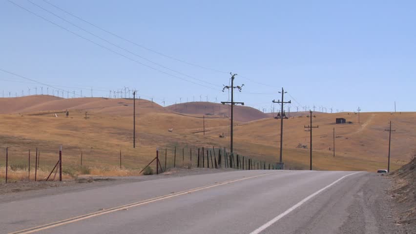 Stationary view of power-generating windmills in the Altamont Pass area of California, near the San Francisco Bay Area. A number of cars pass from far to near and from right to left. Sunny afternoon. Royalty-Free Stock Footage #3411412551