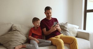 Loving father showing new cool mobile amusing application to little son, sit together on couch staring on smart phone screen, enjoy videocall to family, having fun on-line, spend free time on internet