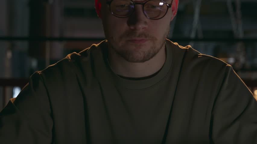 The cyber criminal starts his work, opens a laptop in a dark room in close-up. A 30s year old man wearing glasses and a T-shirt. The laptop screen is displayed in the glasses screen. High quality 4k Royalty-Free Stock Footage #3411688913