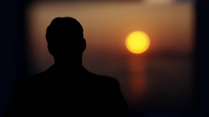 A man stands in a room and looks out the window at the setting sun. Royalty-Free Stock Footage #3411915039