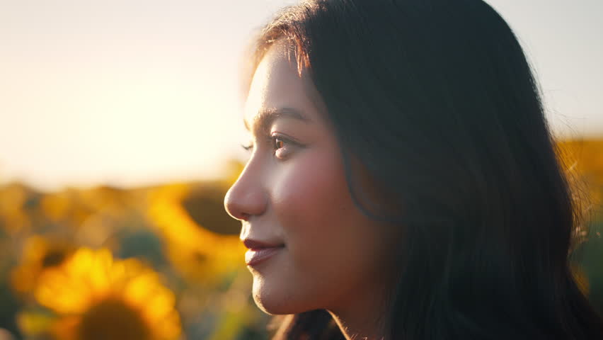 Closeup face of Asian woman opening eyes on sunset background. Young female looking straight at the sunflower field landscape.