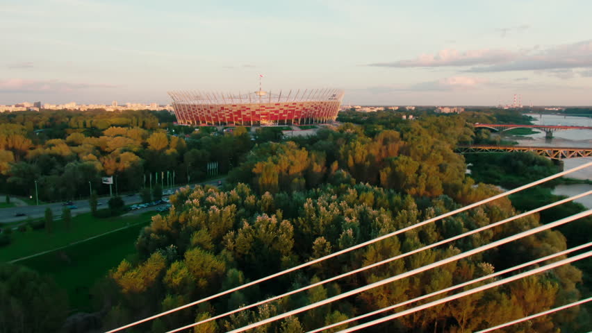 Aerial view of Stadiumin Warsaw, Poland. Roof Football arena with red and white facade. Bridge and Vistula River. 4K zoom out shot