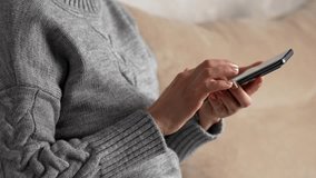 Woman using smartphone and sitting on sofa, side view. Woman hand with cell phone touching screen, close-up 4k
