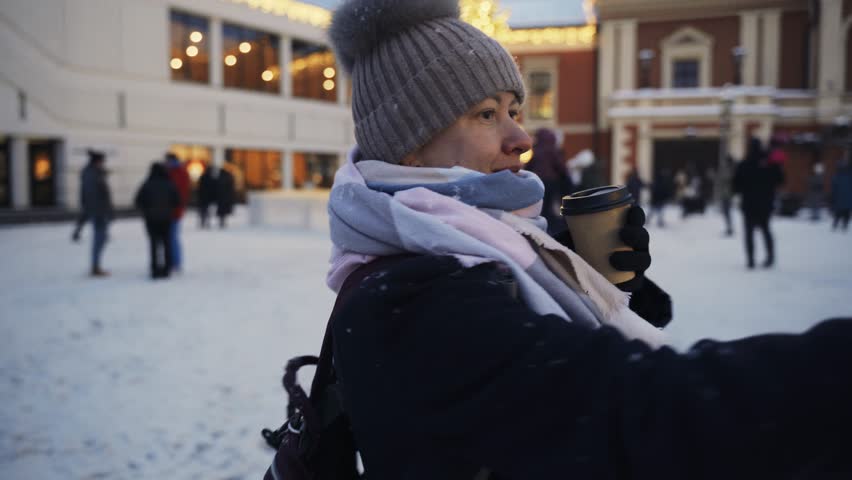 A woman walks through a winter city, shoots video on a smartphone and drinks coffee.