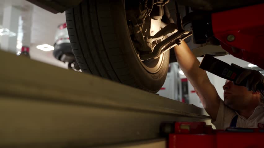 Auto mechanic uses hydraulic lift for undercarriage check. Car suspension inspection at workshop. Vehicle maintenance, steering system diagnostics. Skilled tech servicing auto, equipment use. Royalty-Free Stock Footage #3412226691