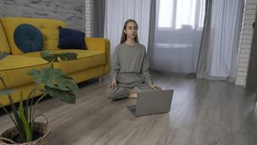 Woman who sits on the living room floor in front of a laptop and meditates online