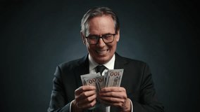 A man in a business suit with a tie joyfully counts dollars in his hands, then displays them on camera. He is extremely happy holding the money. He is on a dark gray background. Camera 8K RAW.