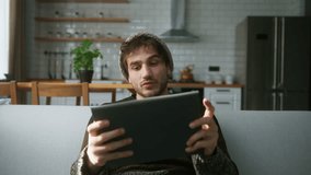 Young man with beanie sitting on sofa at home with kitchen background, playing excited concentrated engrossed in interesting mobil game with tablet computer