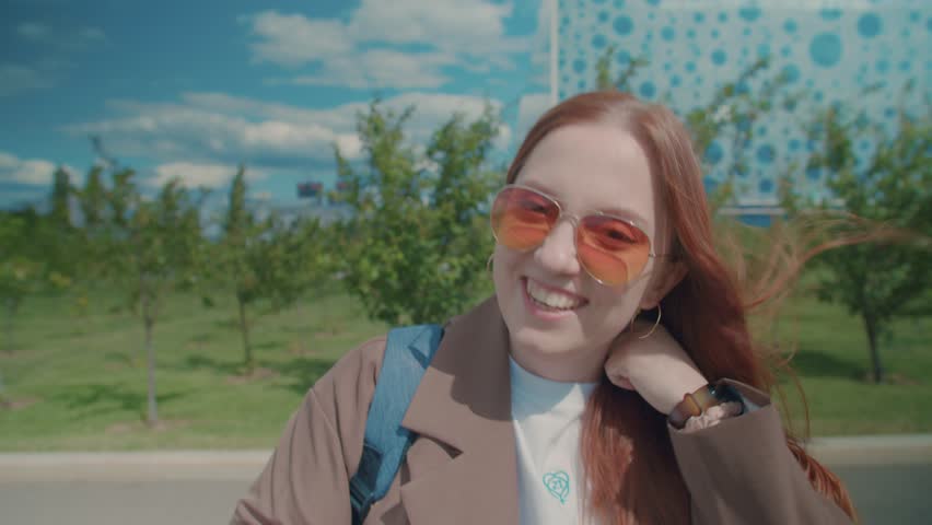 A red-haired girl in sunglasses and a white T-shirt walks along the road along the apple trees.