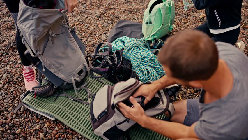 Shot from above, a brunette guy in a gray T-shirt takes out special equipment and insurance for rock climbing from his backpack, along with his two girlfriends who also disassemble their backpacks and