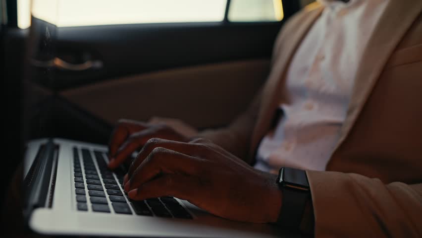 Close-up shot of a man Businessman with Black skin color and in a brown jacket works and types on a laptop while driving in the passenger seat in a modern car interior
