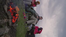 Diverse group of tourists resting in camp. Hiking buddies sit near campfire and talk. Hikers in campsite in mountains. Travelers during their vacation trip. Nature discovery and tourism concept.