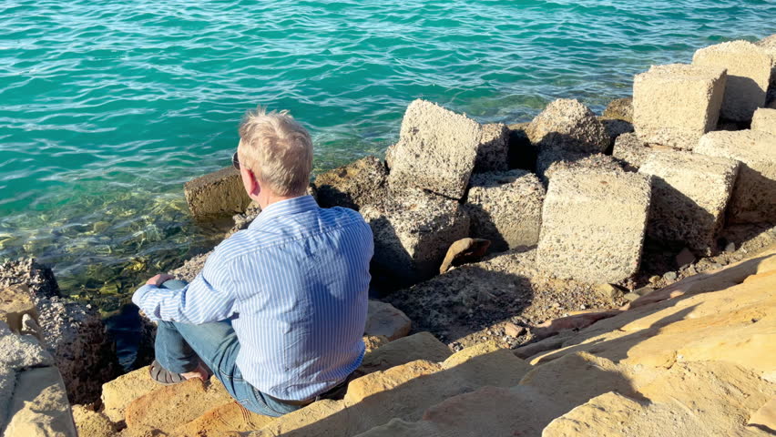 Mature Caucasian man sits alone on rocky shore by the sea.