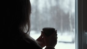 Long hair woman standing among the window and drinking hot tea from mug. Winter landscape. Lady drinks coffee looking at snowfall. Depression seasonal affective disorder female sad alone. Flu season - Powered by Shutterstock - Get 15% off with code: PIKWIZARD15