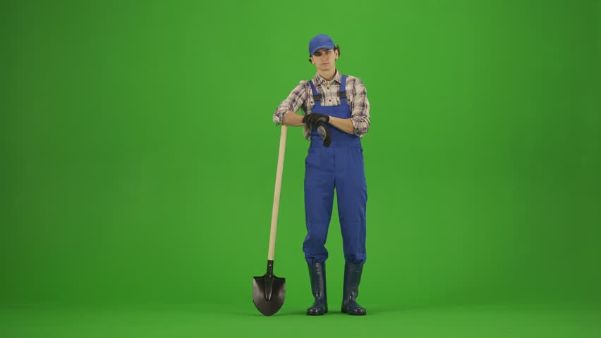 Portrait of male in overalls and rubber boots on chroma key green screen. Man gardener stands leaning on a shovel and smiles showing thumbs up.