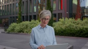 Mature gray-haired woman Caucasian female senior lady greet client online talking laptop device business communication on computer smiling happy middle-aged businesswoman distant call in city outdoors