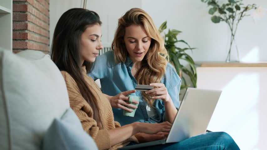 Video of  of two happy young women using computer while paying with credit card their vacation sitting on the couch at home.