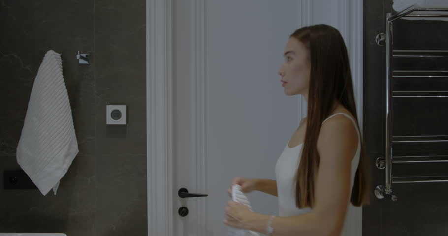 Beautiful woman in the bathroom, getting ready to shower. Young woman hangs a towel and looks in the mirror. Morning routine. Modern home