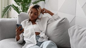 relaxed young black girl in headphones watching movie or film on streaming service on her smartphone while lying on the sofa