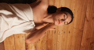 Vertical clip. Young woman in towel relaxing in wooden sauna at spa. Asian woman in bathrobe doing body treatment in sauna.