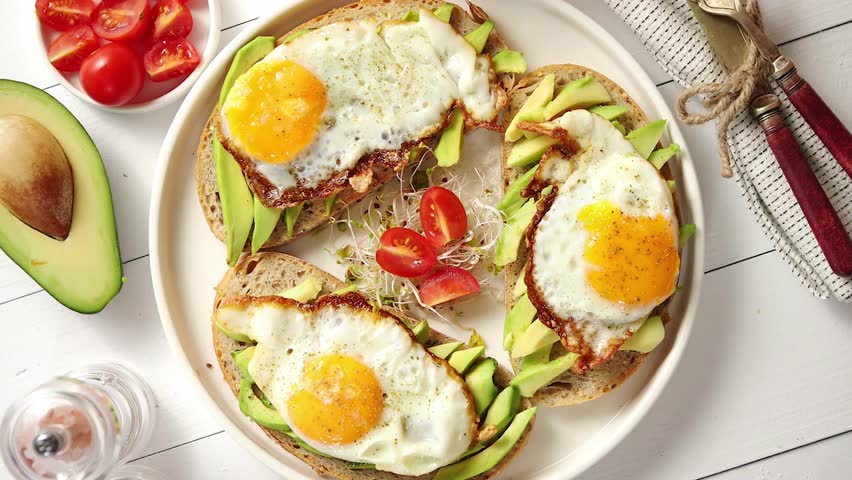 Delicious healthy breakfast with sliced avocado sandwiches with fried egg on top of bread. With orange juice, cherry tomatoes, radish sprouts, salt and pepper. Royalty-Free Stock Footage #3413379357
