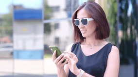 Smiling young woman in sunglasses using smartphone typing text answering messages chatting online looking mobile screen social media app. Girl is outdoor urban sunset city park holding phone in hands