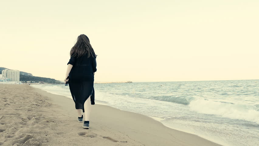 Slow motion of woman in black long waving scarf walking on the beack