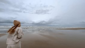 a girl in woolen clothes walks along a wide beach near the northern sea. enjoying nature, listening to the sound of the sea, the north wind blowing on her develops her hair. High quality video