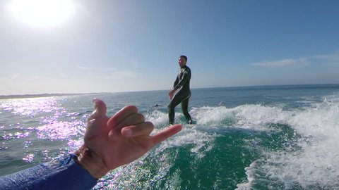SLOW MOTION LENS FLARE POV Stoked cameraman giving shaka sign to young surfer successfully riding a wave. Surfboarder riding a wave and jumping into water as man behind camera congratulates his friend