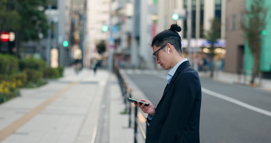 Phone, networking and Japanese businessman in the city reading company email on technology. Career, travel and professional young male person scroll or browse on cellphone with earbuds in urban town.