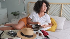 Young multiracial latina sitting on bed checking packing list before summer holidays. Preparing suitcase for vacations. 4k resolution video. Vacation concept.