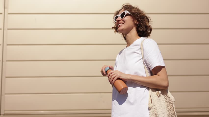 Cheerful millennial young woman opens a reusable metal bottle and drinks fresh water on sunny summer day outdoors on modern building wall background. Zero waste, eco-friendly, no plastic concept. Royalty-Free Stock Footage #3413595293
