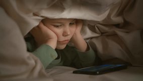 A boy in bed is watching something interesting on the phone. Children's bedtime stories, on the phone. Children's health, sleep disorders, vision loss.