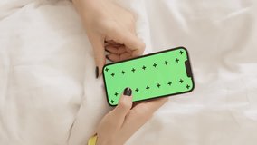 Vertical video. Concept of Ordering Food or Room Cleaning in a Hotel. Female Hands Scroll Through a Smartphone with a Green Screen Mockup and Tracking Markers While Lying on the Bed. Top View.
