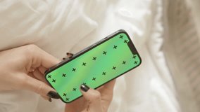 Vertical video. The Camera Moves Around a Woman on the Bed Scrolling Through a Green-Screen Smartphone. Hotel Concept, Room Service Order. with chroma key green screen