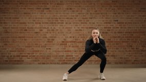 Side lunges. A trainer skillfully demonstrates how to perform the exercise, side lunges. A girl in good physical shape. She is in black sportswear in a loft gym with a brick wall. Camera 8K RAW. - Powered by Shutterstock - Get 15% off with code: PIKWIZARD15