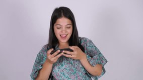 A young Indian woman playing a game on her smartphone - white background, mobile entertainment. Excited young Asian female playing a video game on handheld gadget - cellphone, game and streaming, d...