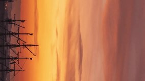 Vertical video. Masts of Old Sailing Ships and a Torn Flag Fluttering in the Wind atop them. Time-Lapse of the Rising Sun and Fiery-Colored Clouds.