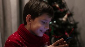 Christmas atmosphere in the house, a 9-year-old Caucasian boy with a phone reads and writes congratulations on the smartphone screen. boy wishes his family Merry Christmas