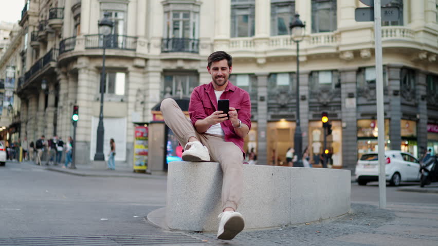 Focused man in casual wear using smartphone app on city bench, urban life and digital engagement. blurred cityscape underscores integration of technology in everyday routines, scrolling social media Royalty-Free Stock Footage #3413817791