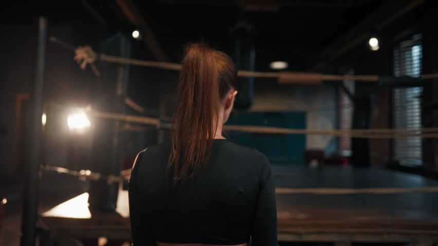 Barefooted woman boxer fighter in sportswear with ponytail going to boxing ring between ropes in boxing gym. Girl preparing to her training, workout. Professional sport, competitions, exercising.